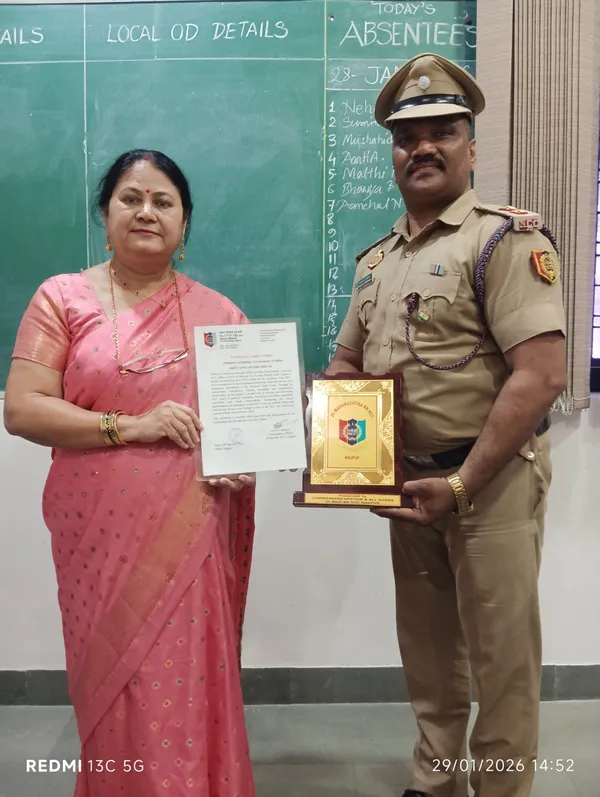A woman in a pink saree and a man in a khaki uniform stand in front of a green chalkboard, holding a framed certificate and a golden plaque.