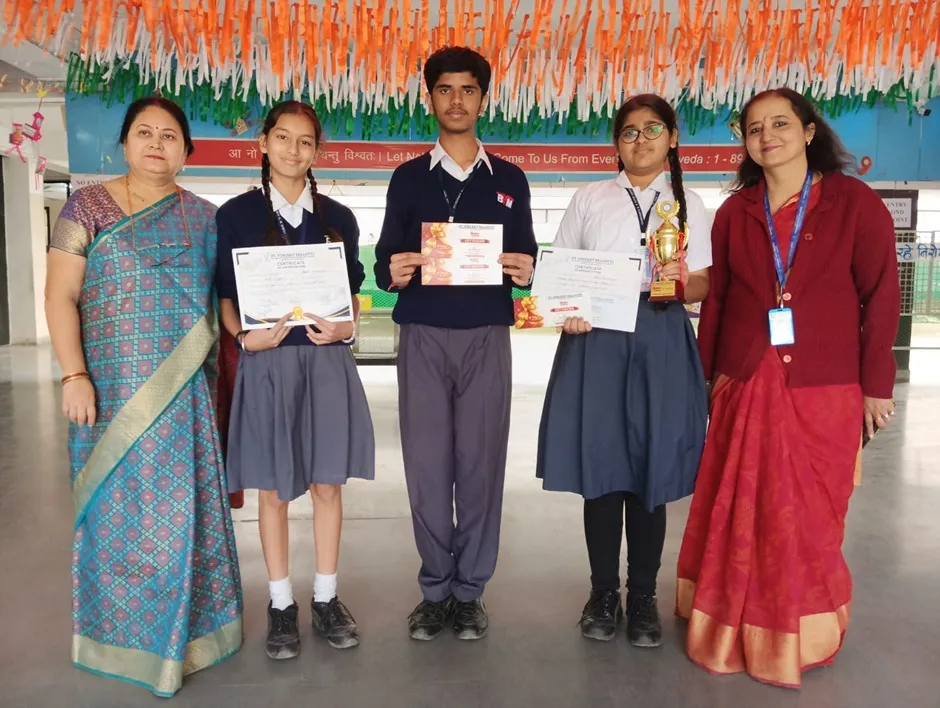 Group photo of three students holding certificates and a trophy, standing between two women teachers in a hall decorated with tricolour streamers. Bhavan's BVM Ashti.