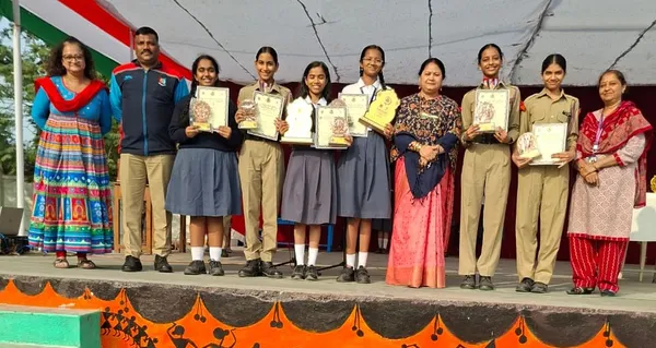 Group photo on a stage of several girl students in uniform and four adults holding certificates and trophies under a tent, Bhavan's BVM Ashti.
