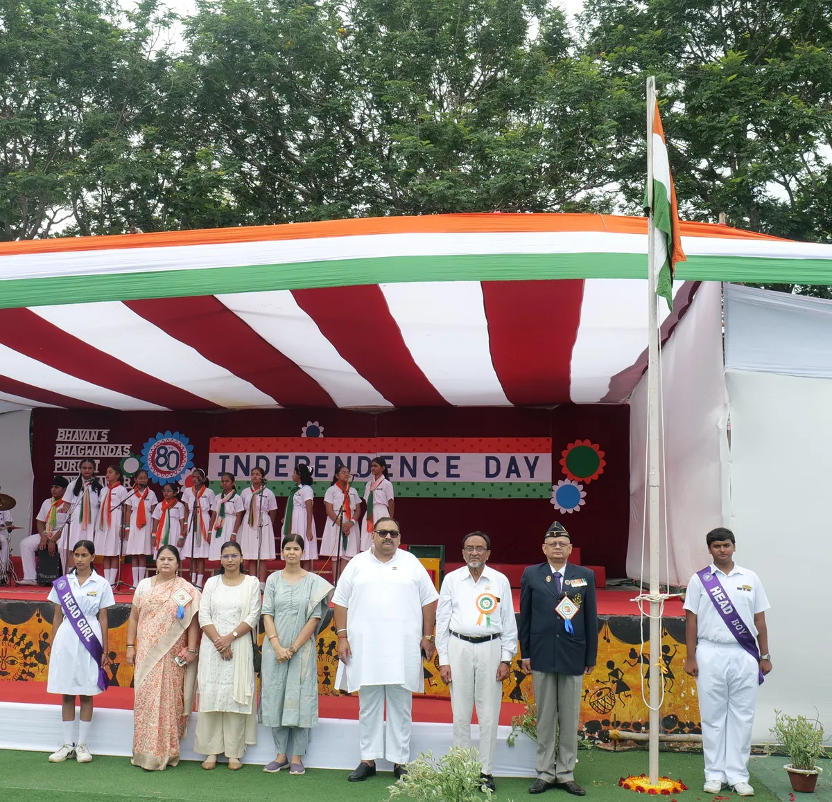 Officials, guests and head students standing by a flagpole in front of a decorated Independence Day stage, with students in white uniforms standing on the stage behind them at Bhavan's BVM Ashti.