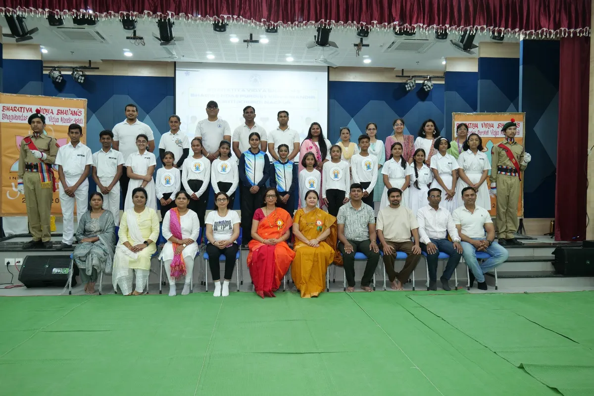 Group photo of students, teachers and guests on a stage with two uniformed cadets at the sides, banners and a screen behind them at Bhavan's BVM Ashti.