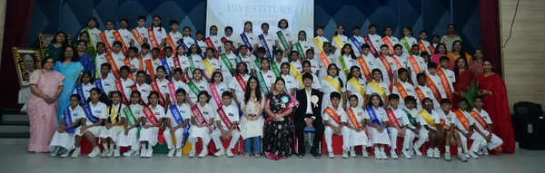 Wide group photo of many students in white uniforms wearing colored sashes, with several adult women, posed in front of a stage backdrop. - Bhavan's BVM Ashti