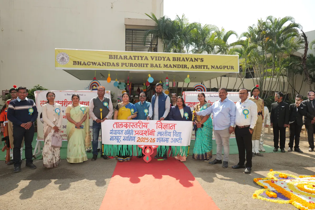 Group photo of men and women wearing badges, standing on a red carpet holding a banner in front of Bhavan's BVM Ashti school building, with a flower rangoli in the corner