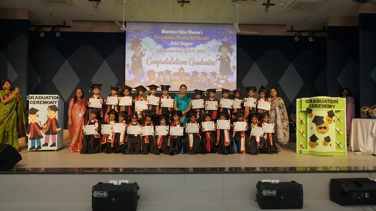 Group photo on stage of young children in graduation gowns and caps holding certificates, with teachers standing alongside and graduation ceremony decorations and congratulations screen behind them.