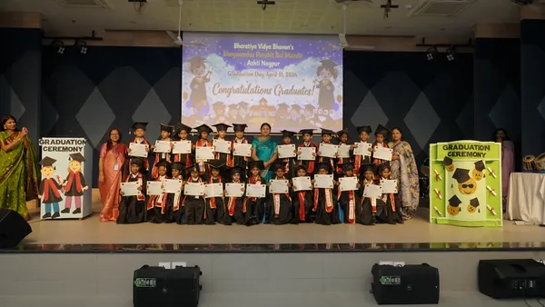 Group photo on stage of young children in graduation gowns and caps holding certificates, with teachers standing alongside and graduation ceremony decorations and congratulations screen behind them.