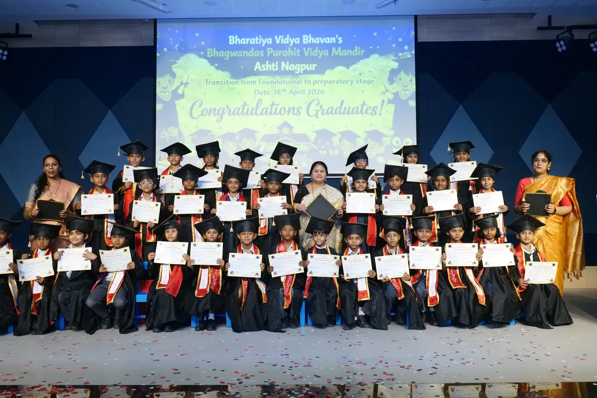 Group photo of young children in graduation caps and gowns holding certificates, with three women standing among them in front of a stage screen, at Bhavan's BVM Ashti.