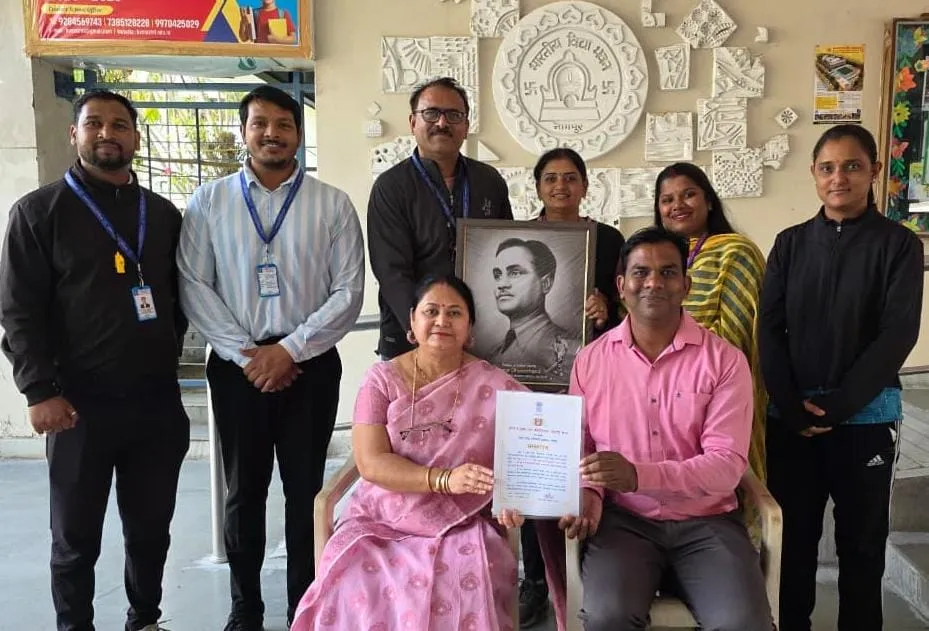 Group photo of eight people, two seated holding a framed certificate, with a framed black-and-white portrait held behind them, posed indoors in front of a wall with decorative relief panels. - Bhavan's BVM Ashti