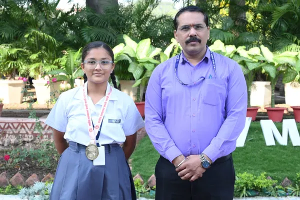 Nishka Dabde wearing a medal standing beside a man in a purple shirt in front of green plants