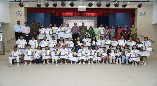 Group photo of students and adults posing with certificates and plaques in a hall with a red curtain at Bhavan's BVM SKN.