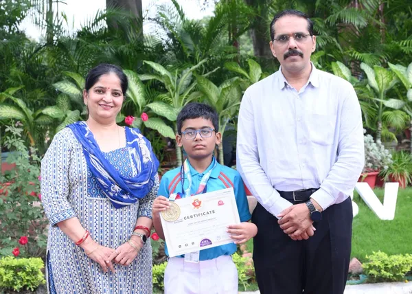 Boy with medal and certificate stands between a woman and a man outdoors in front of green plants