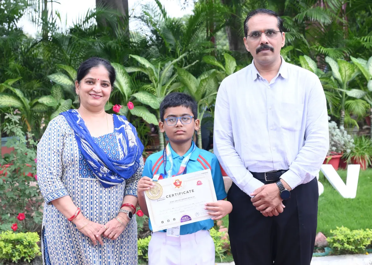 Boy with medal and certificate stands between a woman and a man outdoors in front of green plants