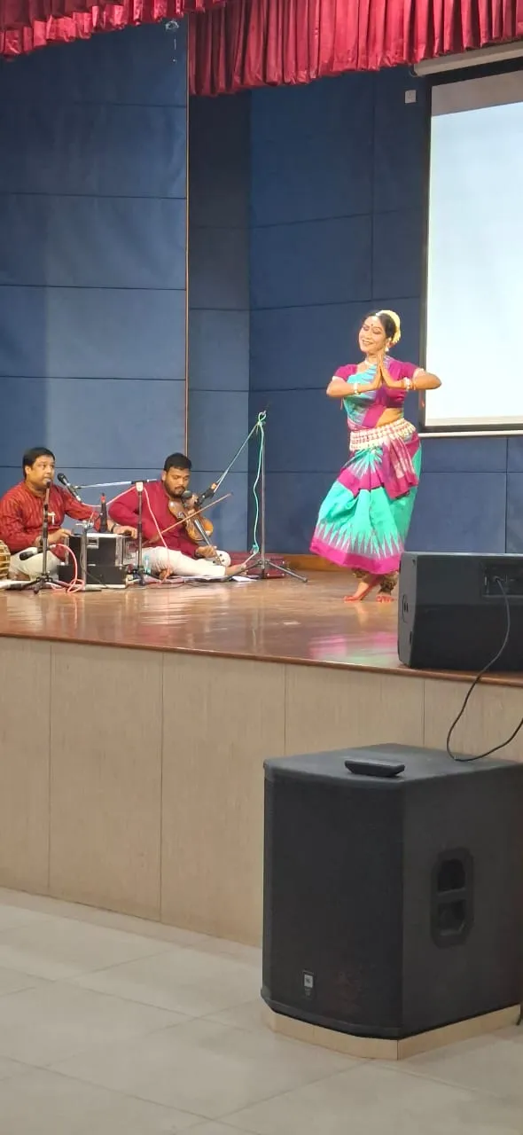 A dancer in a pink and teal costume performs on a stage while two seated musicians play instruments, with speakers in the foreground. Bhavan's BVM SKN.
