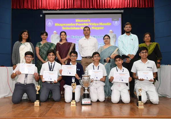 Group photo of six kneeling students holding certificates and trophies with seven adults standing behind them on a stage in front of a projector screen. - Bhavan's BVM SKN