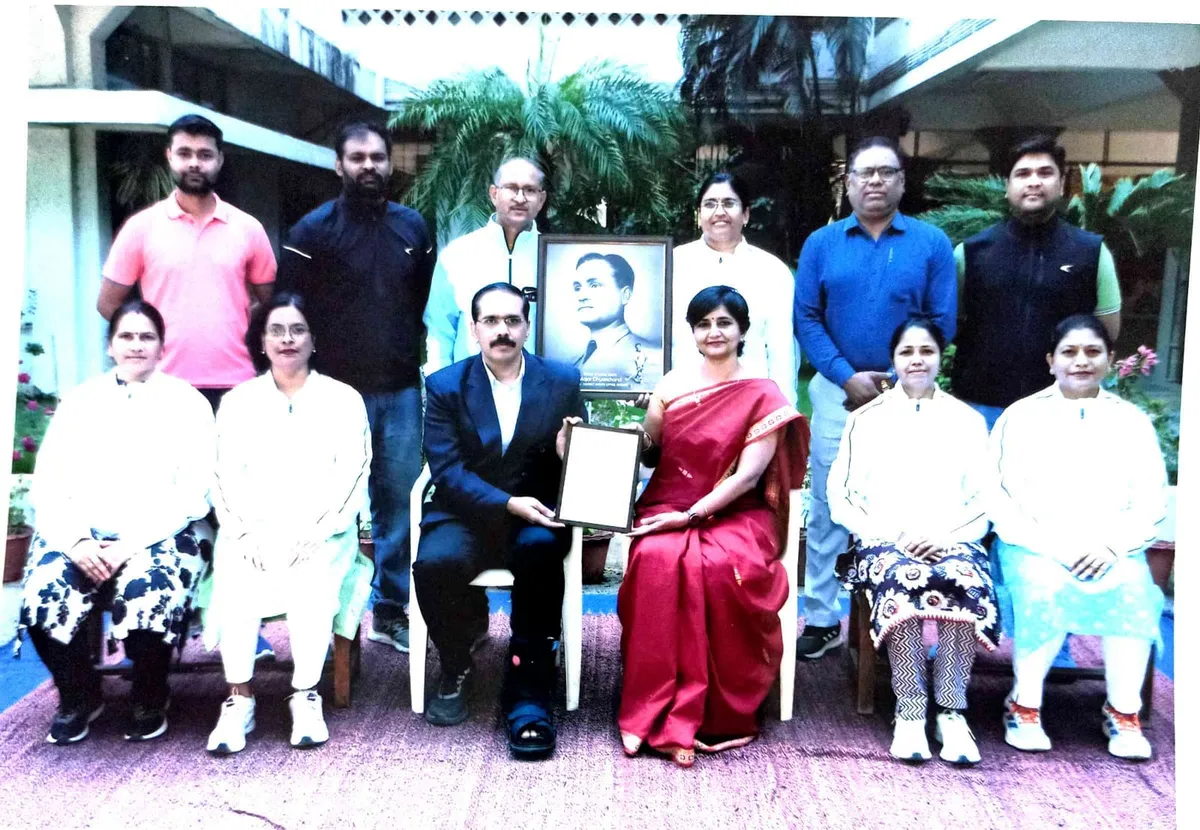 Group photo of eleven adults posed in two rows outdoors, with a man and a woman seated in the center holding a framed item beside a portrait photo at Bhavan's BVM SKN.