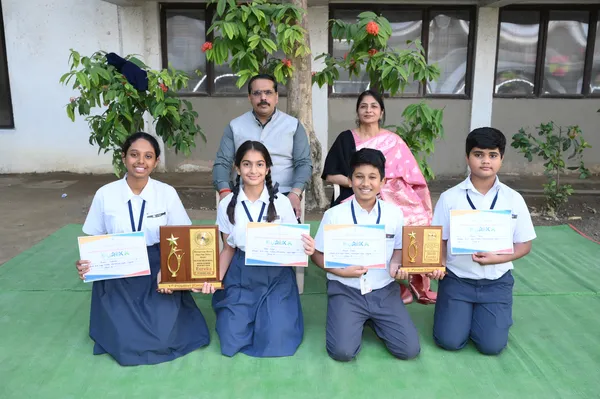 Group photo of four students holding certificates and two plaques, with a man and a woman seated behind them outdoors on a green mat.