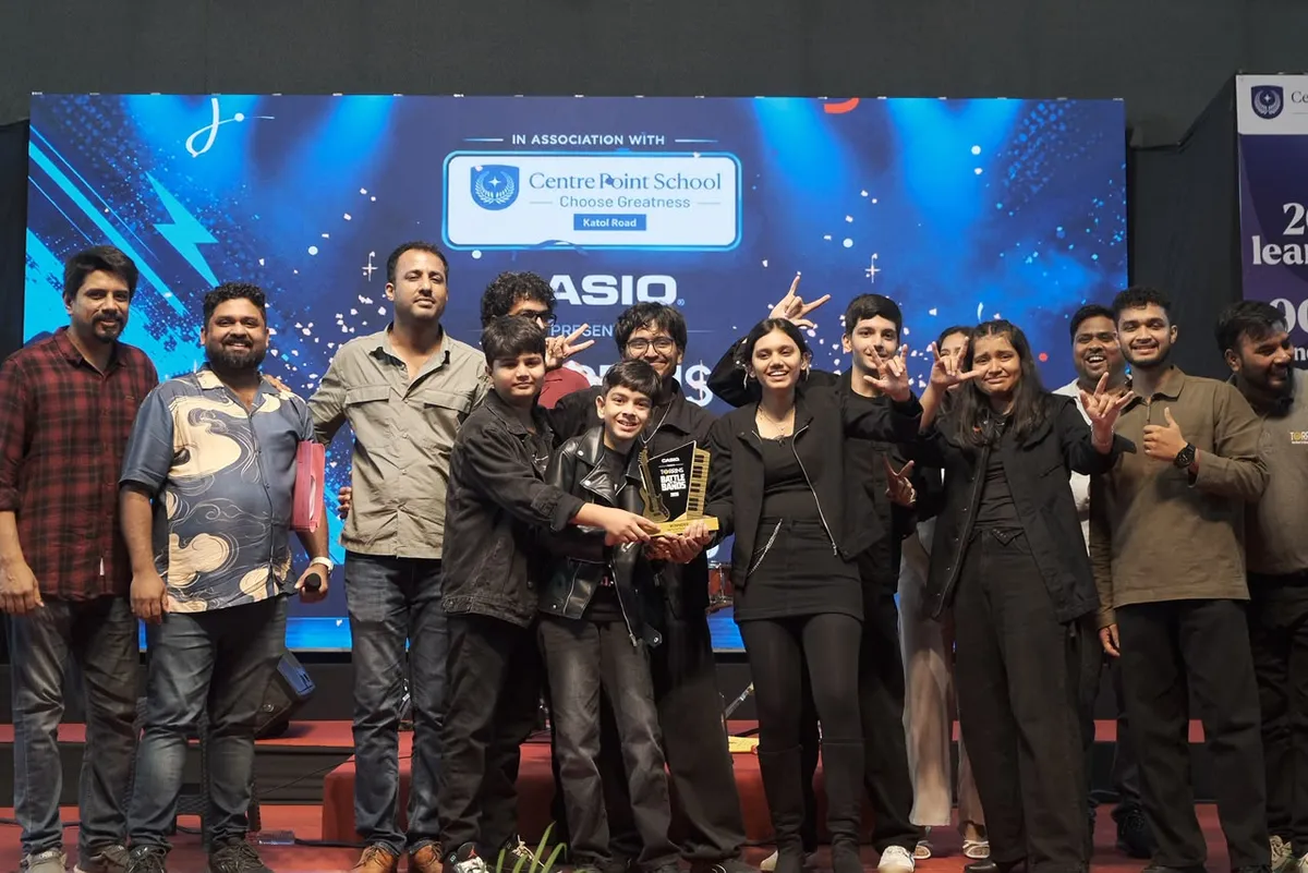 Group photo of about 13 students and adults posing on a stage in front of a screen, with two boys holding a small trophy and several people making hand gestures. - CPS (Katol Road)