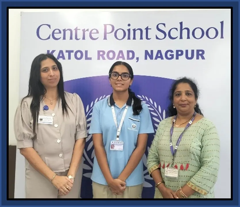 Three people, a student in a blue polo shirt flanked by two women with ID lanyards, posing in front of a school name backdrop.