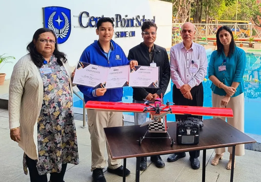Student with three certificates stands with four adults behind a table with a red-winged drone and remote controller, in front of a school sign and a swimming pool
