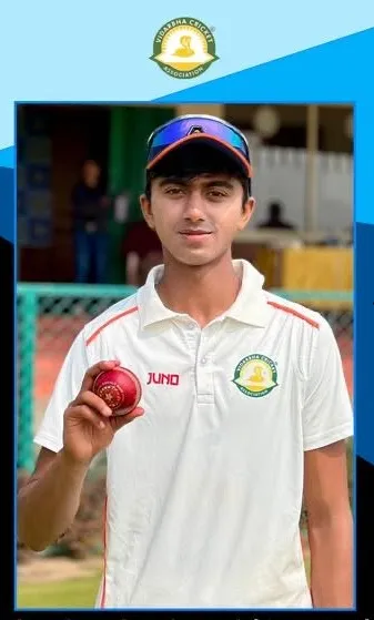 A young cricketer in white jersey and cap with sunglasses holds up a red cricket ball