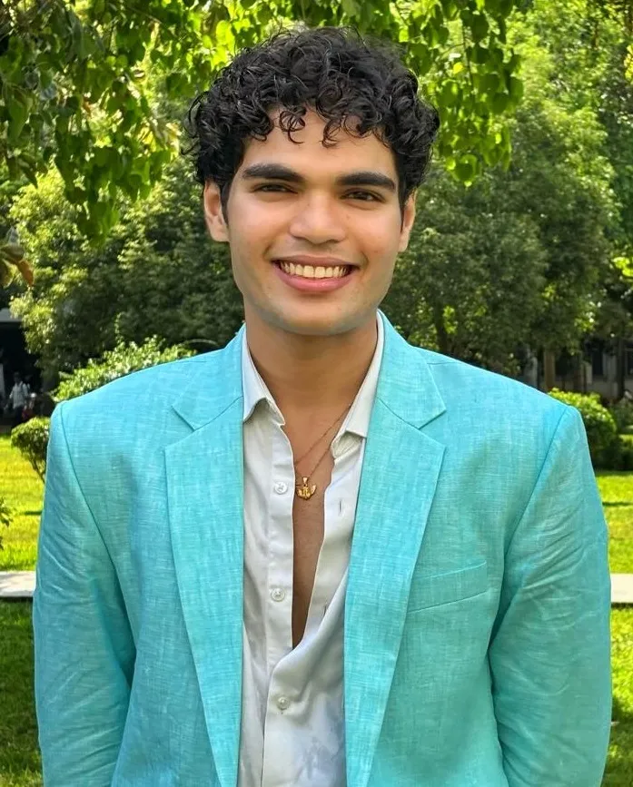 Portrait of Chirag Kubde, a young man with curly dark hair wearing a teal blazer over a white shirt, standing outdoors in front of green trees and grass.