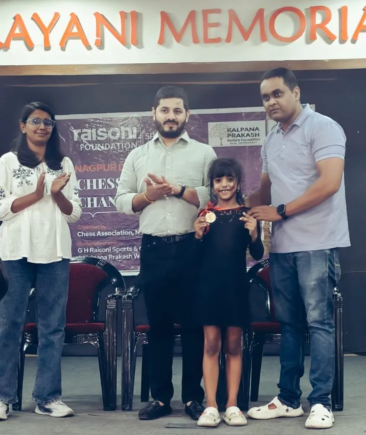 A young girl holding a medal stands on a stage with three adults in front of a chess event banner at CPS (Katol Road).