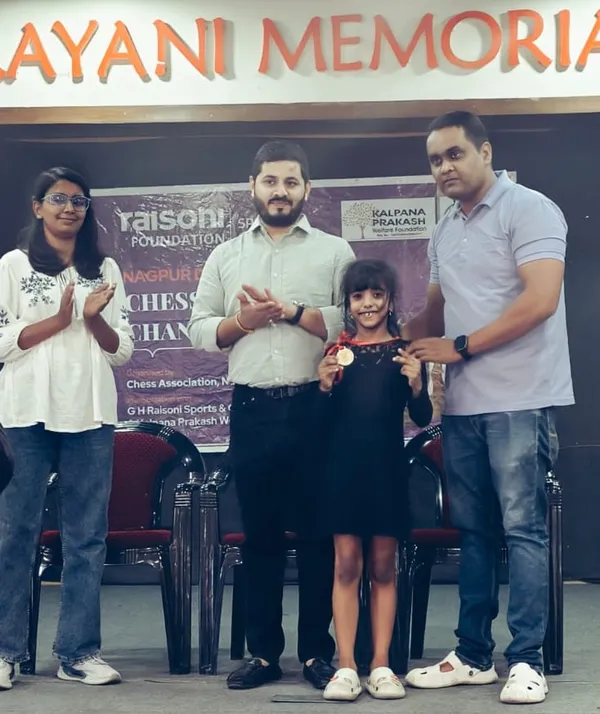 A young girl holding a medal stands on a stage with three adults in front of a chess event banner at CPS (Katol Road).