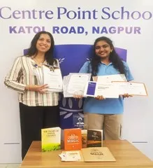 Two women stand in front of a school name backdrop holding certificates, with several books on a table in front of them. - CPS (Katol Road)