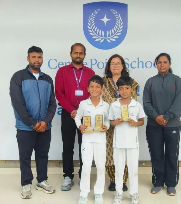 Two boys in white cricket uniforms holding small trophies, standing in front of four adults and a school logo wall.