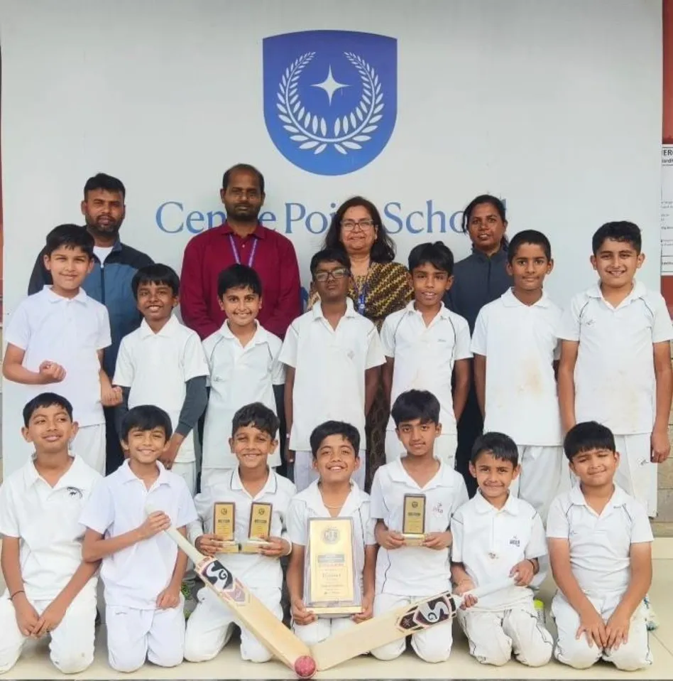 Group photo of young cricket players in white uniforms with trophies and bats, posing with four adults in front of a school logo wall.