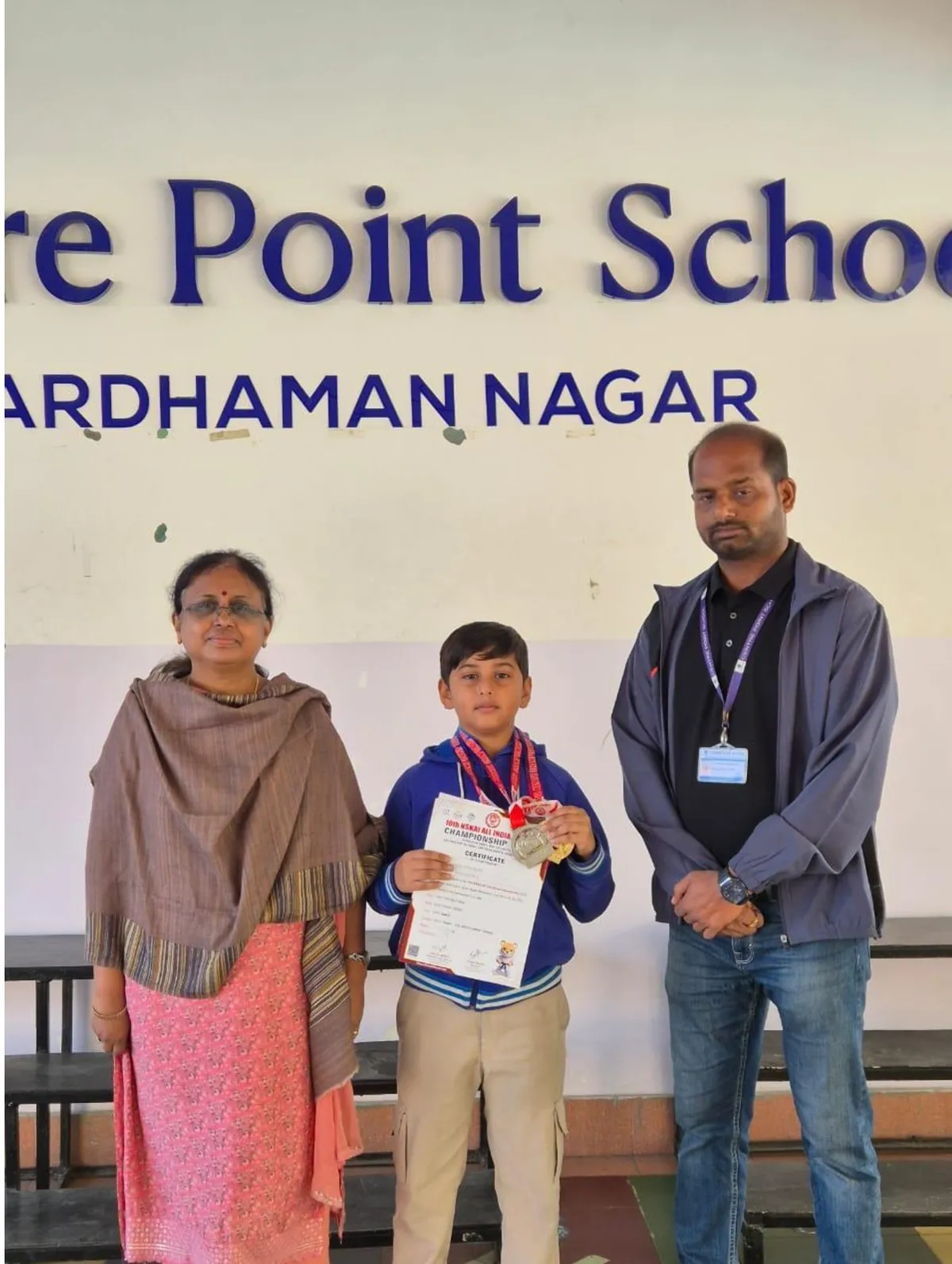 A boy holding a certificate and wearing medals stands between a woman and a man in front of a school name board.