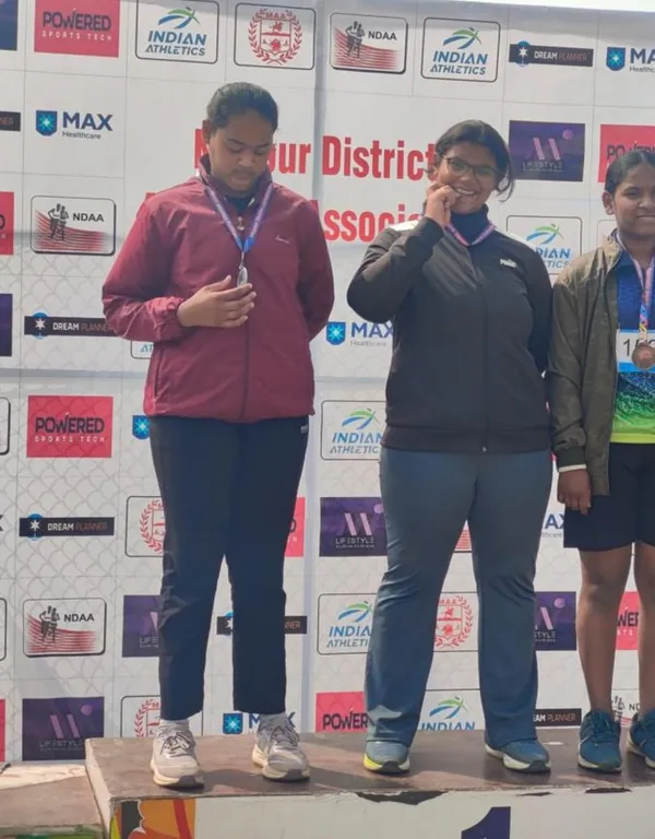 Three girls with medals standing on a podium in front of an athletics sponsor backdrop, one smiling and biting her medal.