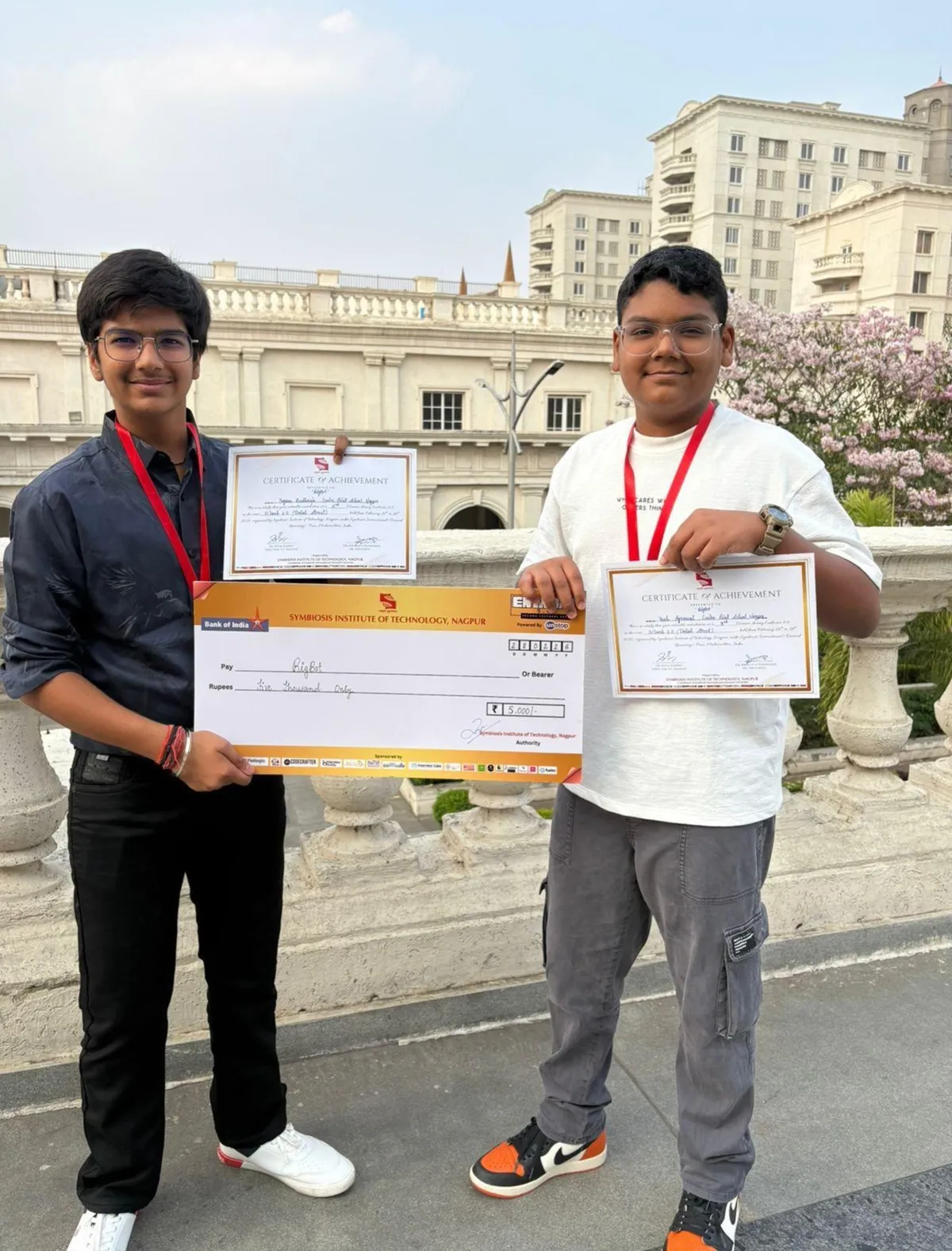 Two boys with lanyards holding two certificates and a large cheque in front of a building with flowering tree.