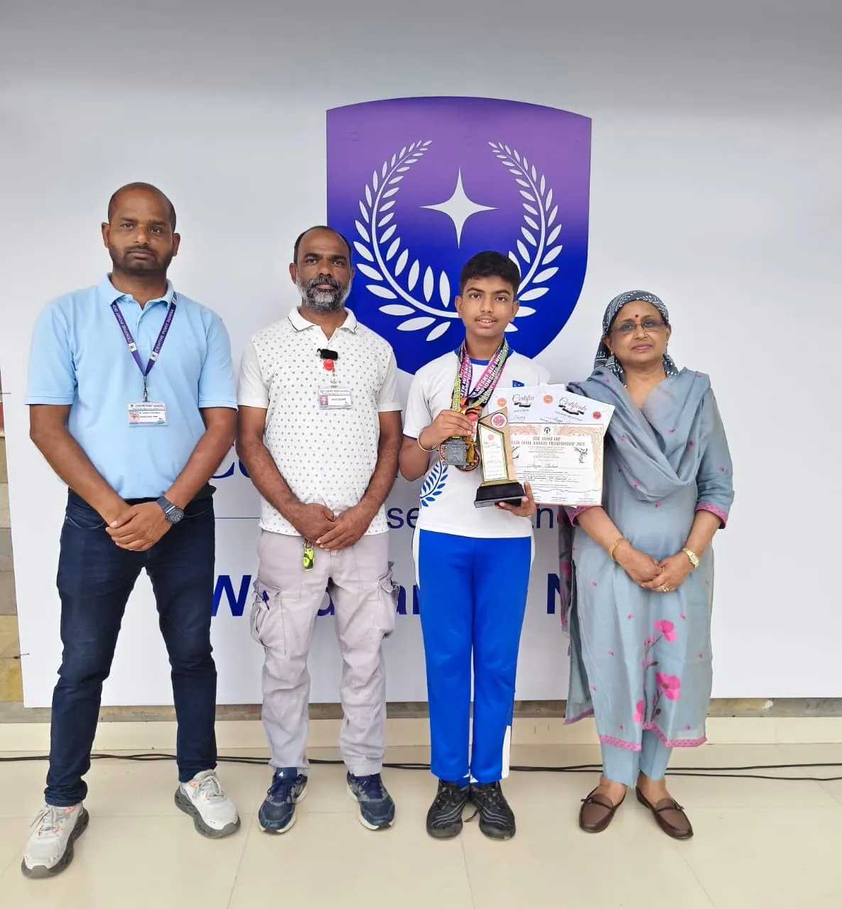 A boy in a white and blue tracksuit holds medals, a trophy and certificates, standing with three adults in front of a wall with a purple emblem.