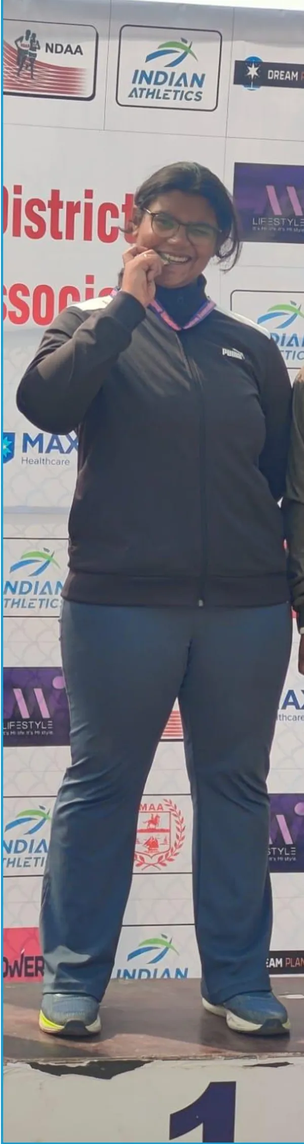 A person in a dark tracksuit stands on a podium in front of a sponsor backdrop, holding a medal at CPS (Wardhaman Nagar).