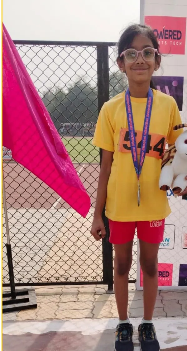 A girl in a yellow shirt with a bib number and a medal around her neck stands in front of a fence holding a soft toy, with a pink flag beside her at CPS (Wardhaman Nagar).
