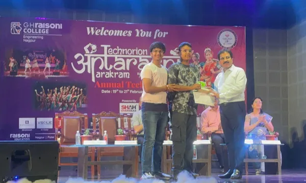 Two students receive an item from a man in a white shirt on a stage in front of an event banner at CPS (Wardhaman Nagar).