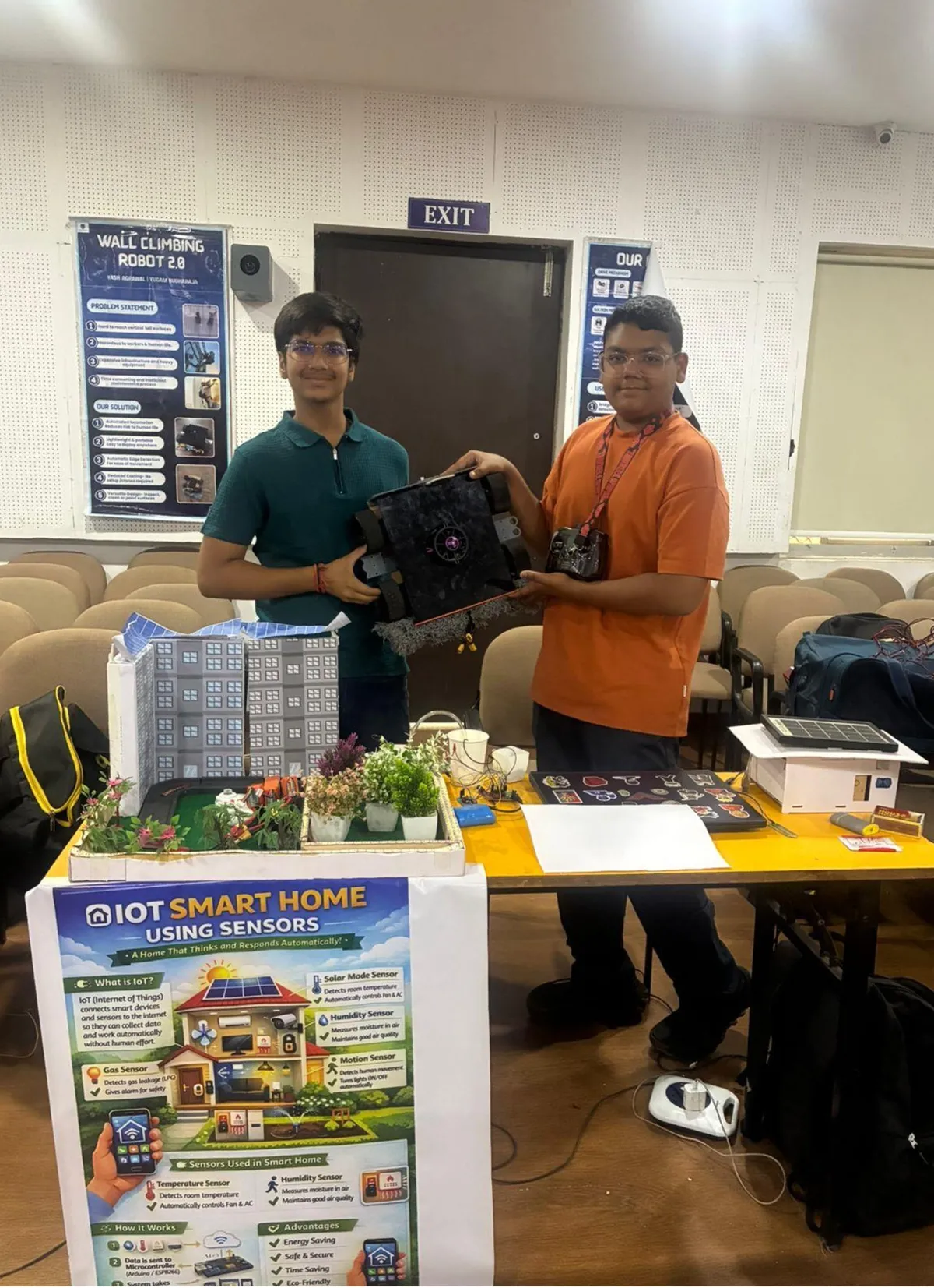 Two students stand behind a table holding a small black robot, with a smart home model and an IoT poster in front of them, at CPS (Wardhaman Nagar).