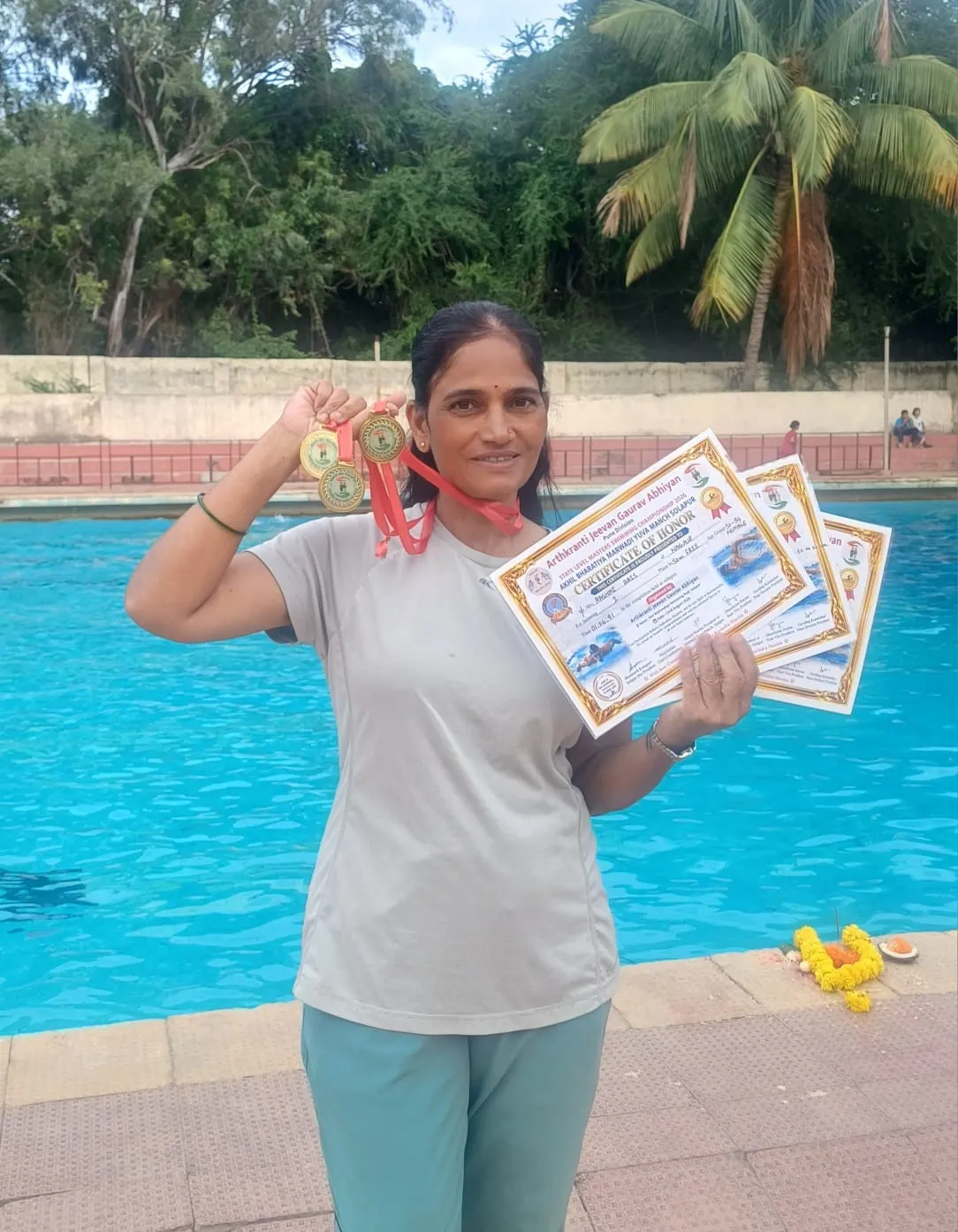 Rashmi Bais stands beside a swimming pool holding up several gold medals and three certificates of honor, smiling at the camera.