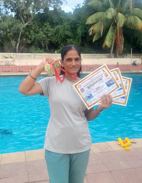 Rashmi Bais stands beside a swimming pool holding up several gold medals and three certificates of honor, smiling at the camera.
