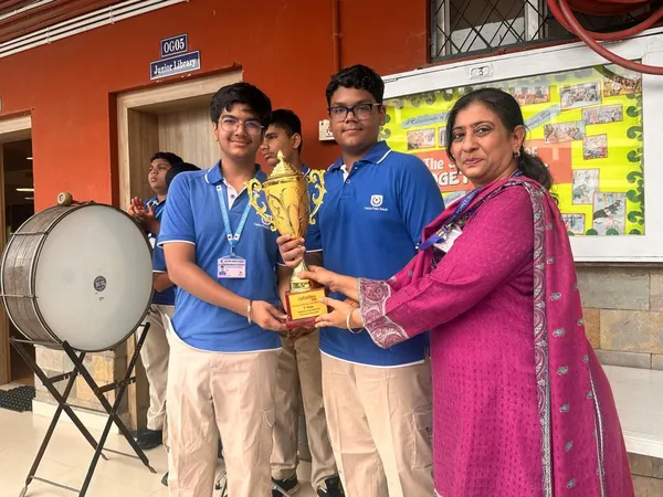 CPS (Wardhaman Nagar): Two students in blue polos and a woman in a magenta outfit hold a golden trophy together in a school corridor with a drum and notice board behind.