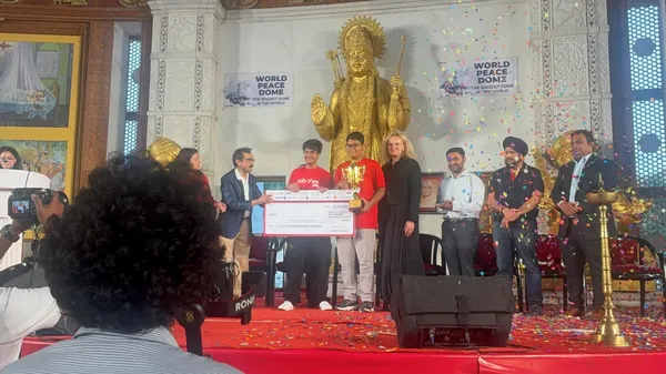 CPS (Wardhaman Nagar): Stage ceremony with two students in red holding a large cheque and trophy among several adults, with confetti, a golden statue and banners behind and a camera operator in the foreground.