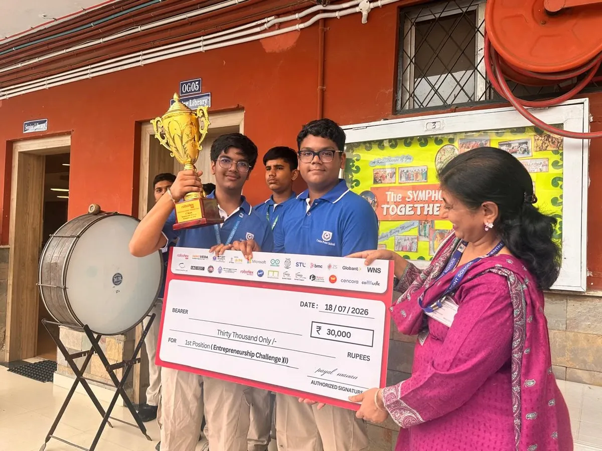 CPS (Wardhaman Nagar): Two students in blue polos hold a golden trophy and a large prize cheque beside a woman in a pink outfit, with other students and a drum behind them.