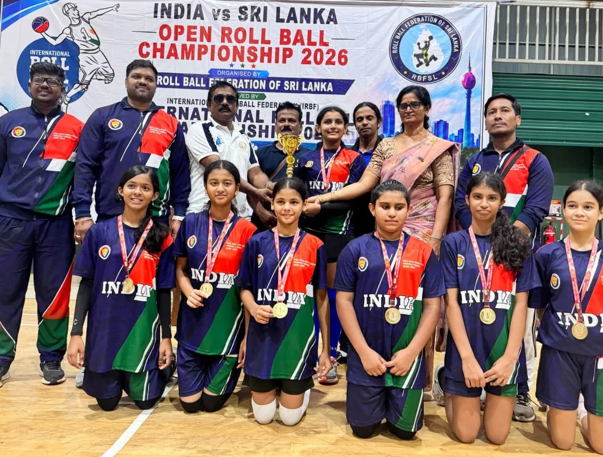Group photo of girls in India jerseys wearing gold medals, with coaches and officials in front of a roll ball championship banner and trophy, CPS (Wardhaman Nagar)