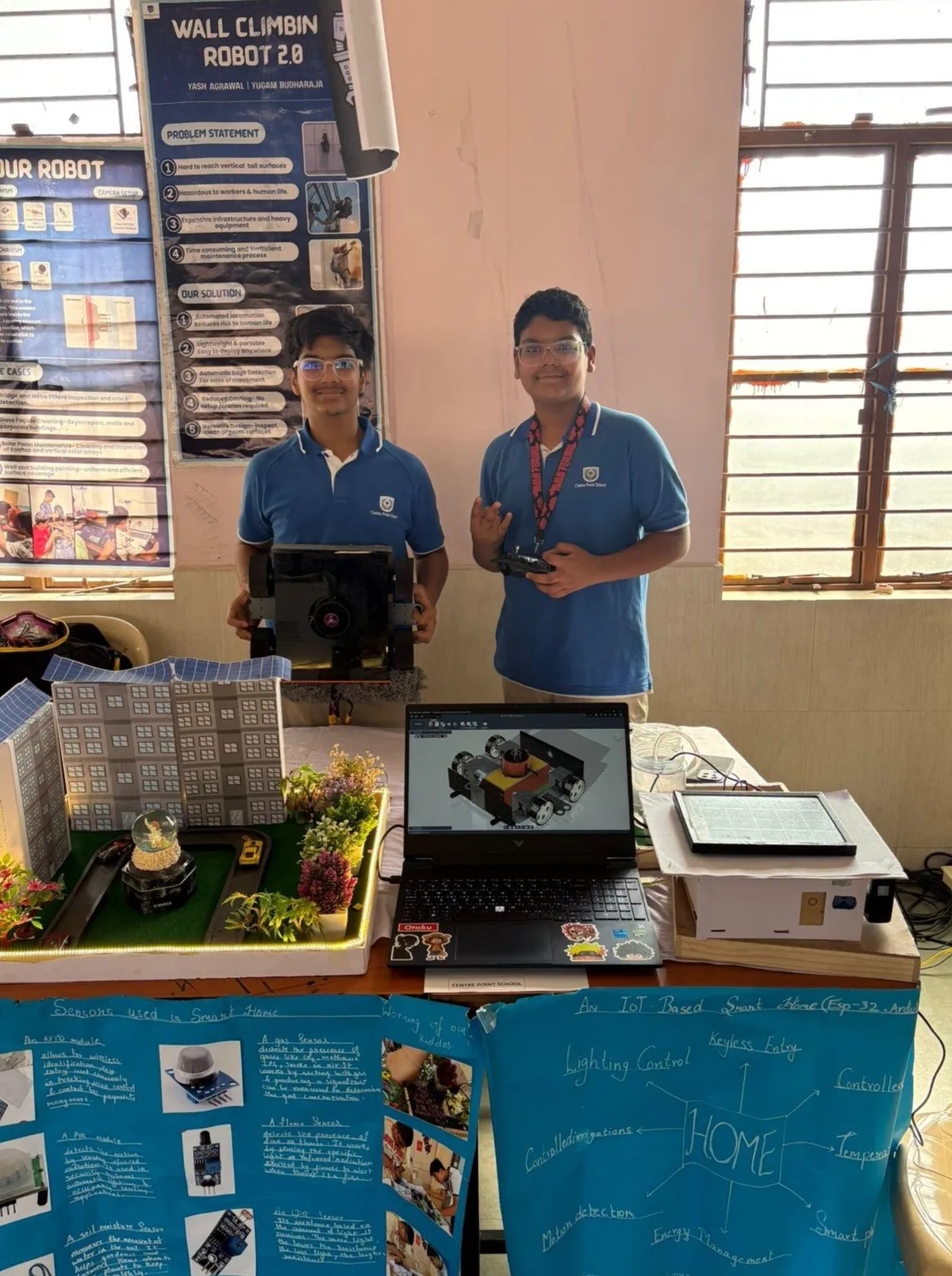 Two students in blue polo shirts stand behind a table with a laptop, a model smart home and a robot, in front of project posters at Skill Fest.