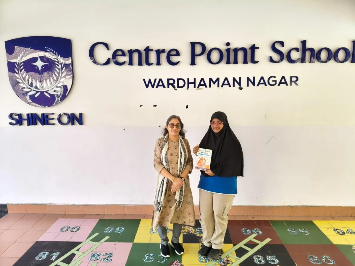 Two people standing under a CPS (Wardhaman Nagar) school name sign on a wall, one holding up a small book, on a colorful tiled floor.