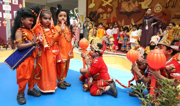Young children in orange and red mythological costumes with crowns and props perform on a blue floor while other costumed children and a teacher look on. - DPS (Kamptee Road)
