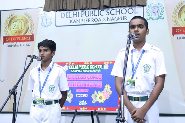 Two students in white school uniforms speaking at microphones on a stage in front of school banners and a decorated board at DPS (Kamptee Road).