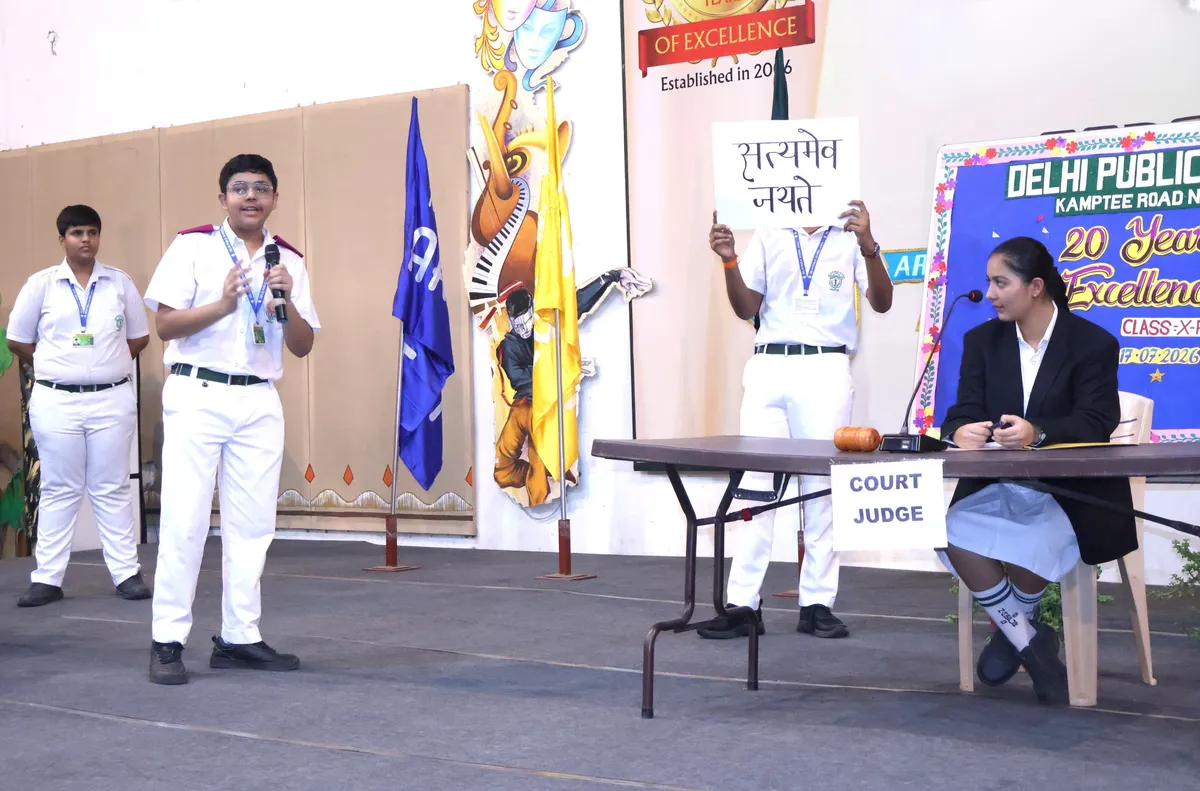 Students of DPS (Kamptee Road) in school uniform perform a skit on stage, one speaking into a microphone, one holding a Hindi sign, and a girl seated at a table labelled court judge.