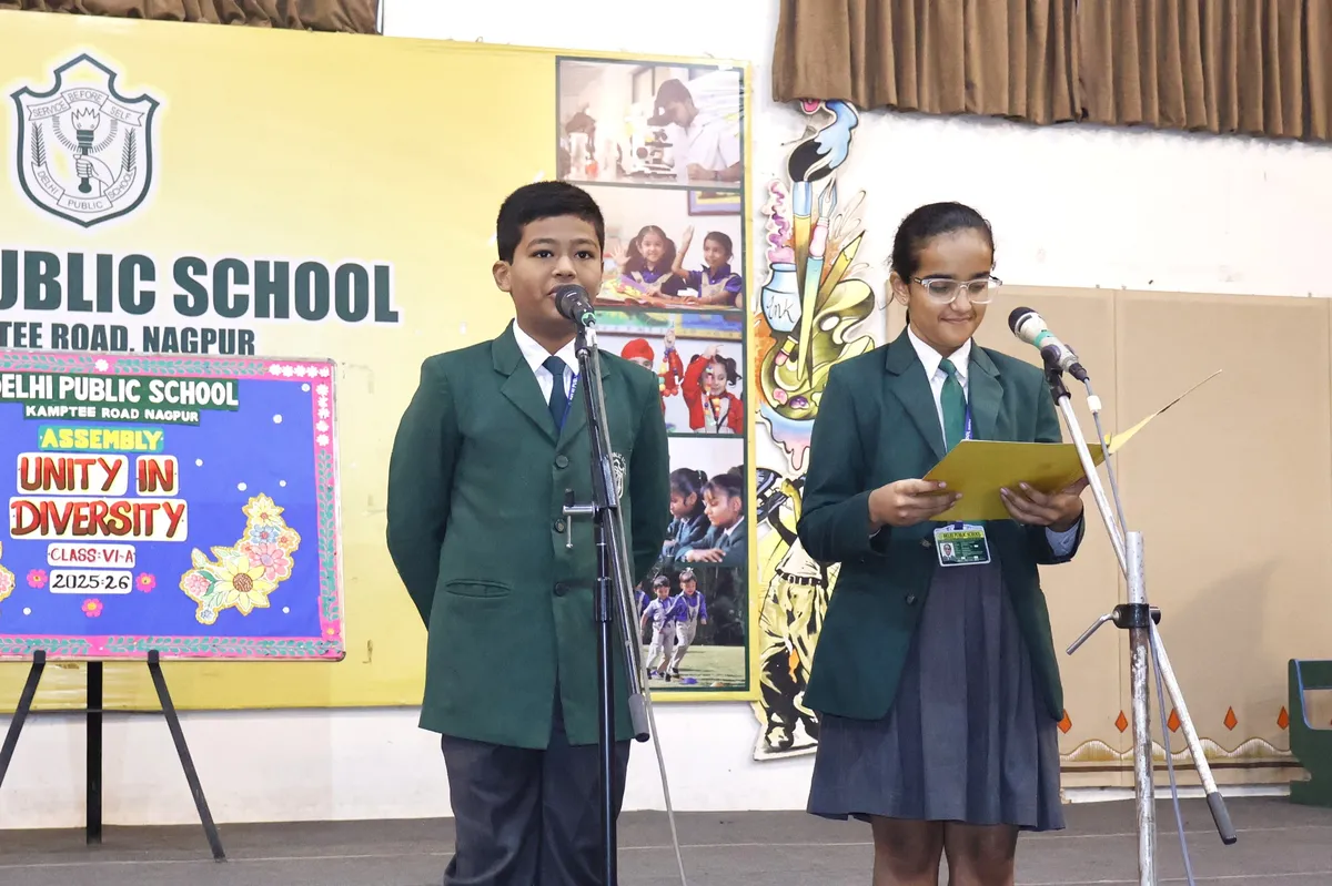 Two students in green blazers speaking at microphones on a school assembly stage at DPS (Kamptee Road), with a decorated board and backdrop banner behind them.