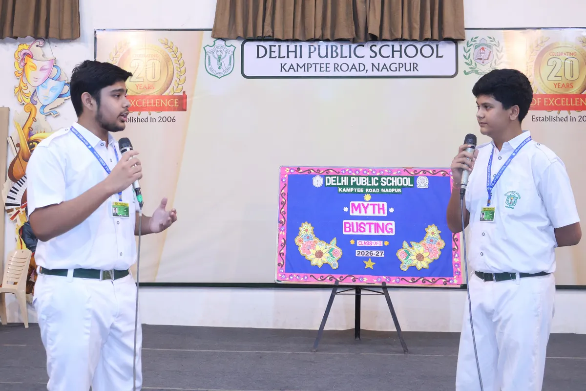 Two students in DPS (Kamptee Road) uniforms speaking on stage with a decorated board and school banner